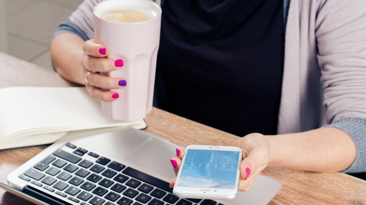 A woman checking her phone and laptop at a desk with coffee